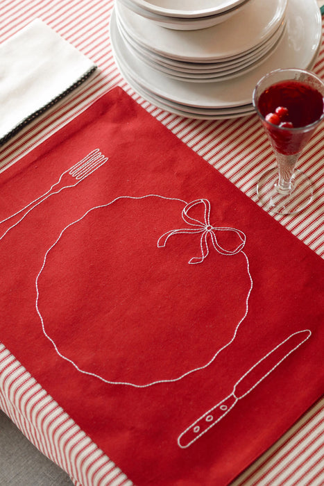Red embroidered placemat on a striped tablecloth beside stacked white plates and a glass of red drink