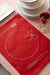 Red embroidered placemat on a striped tablecloth beside stacked white plates and a glass of red drink