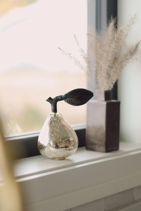 Champagne glass pear on a windowsill beside a tall rectangular vase filled with dried grasses