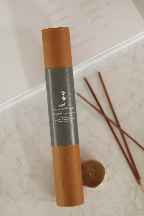 Ember incense tube with a muted green label beside loose sticks and a gold-toned holder on a pale surface