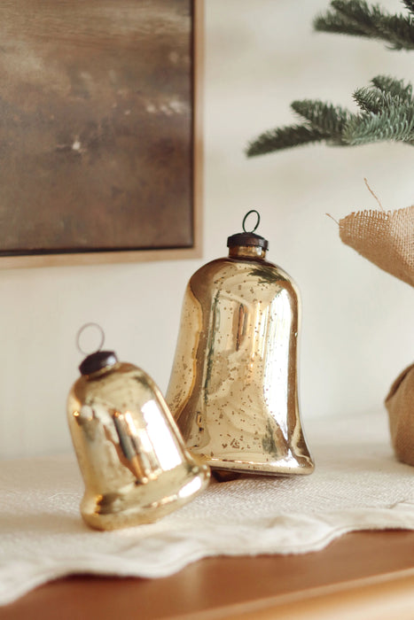 Two gold mercury glass bell ornaments styled on a tabletop beside seasonal greenery
