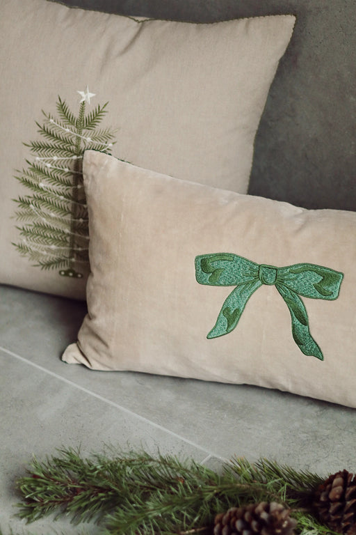 Neutral velvet pillow with an embroidered green bow layered in front of a Christmas tree cushion beside pine branches
