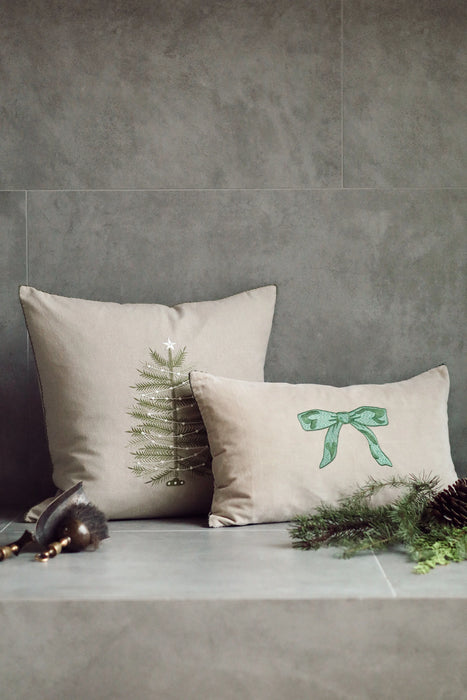 Green bow lumbar pillow beside a square Christmas tree cushion against a grey wall with seasonal greenery