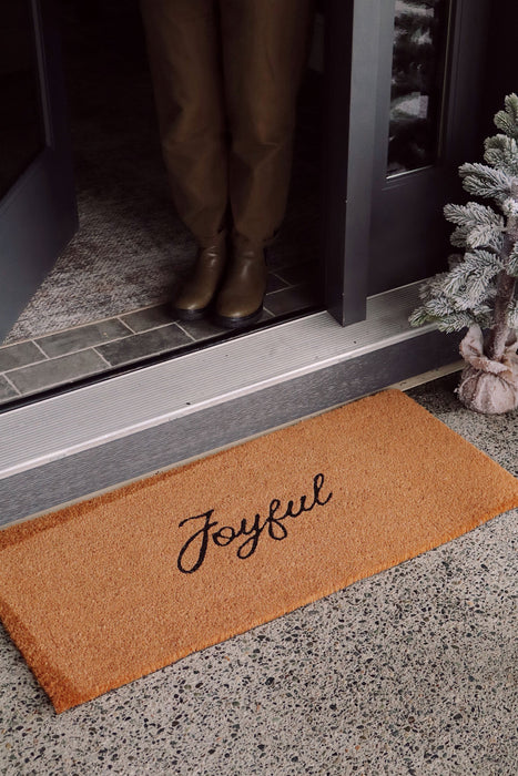Joyful coir doormat at a dark front doorway beside a frosted miniature Christmas tree