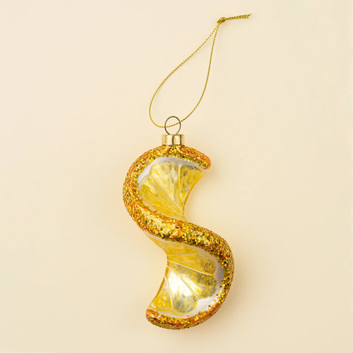 Yellow spiral lemon glass ornament with glittered rind, a gold-tone cap and hanging cord against a cream background