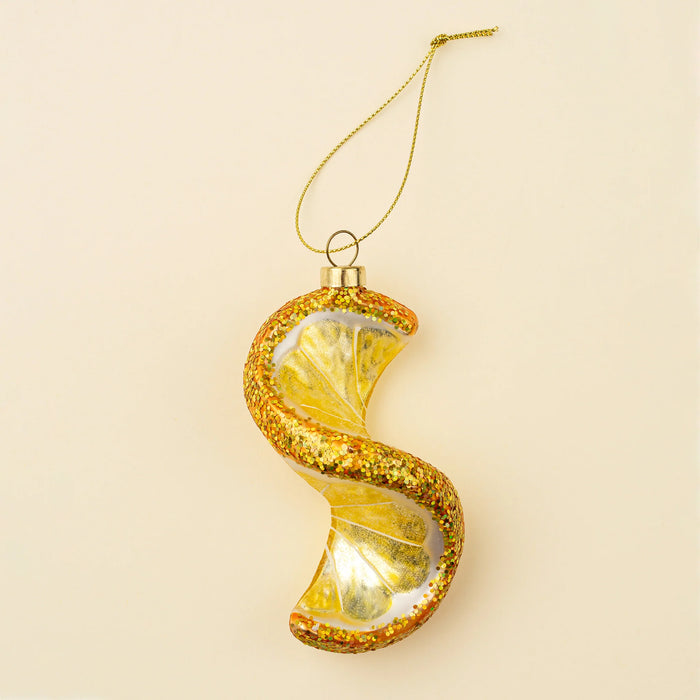 Yellow spiral lemon glass ornament with glittered rind, a gold-tone cap and hanging cord against a cream background