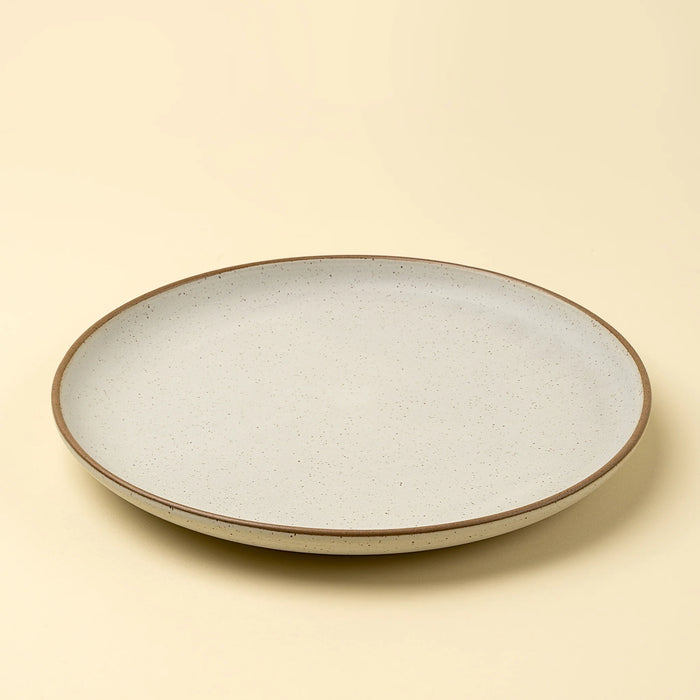 Angled view of an Indaba Rowe ivory stoneware dinner plate with a speckled finish and brown rim