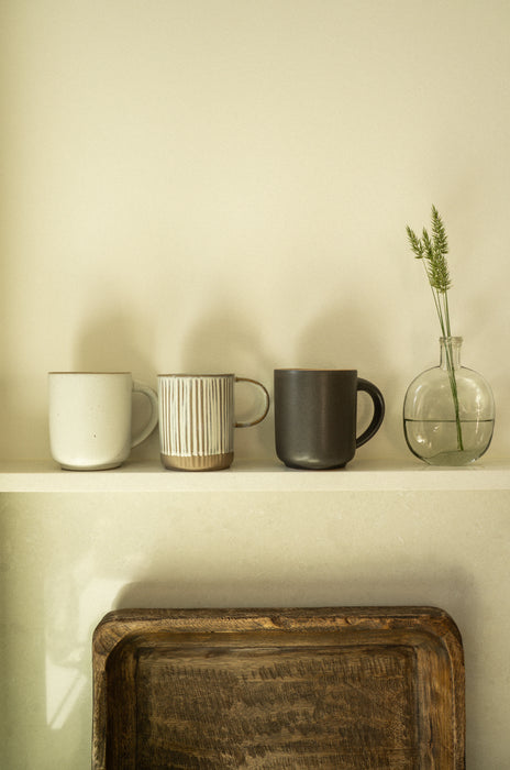 Ivory stoneware mug displayed beside striped and dark mugs and a glass vase on a shelf